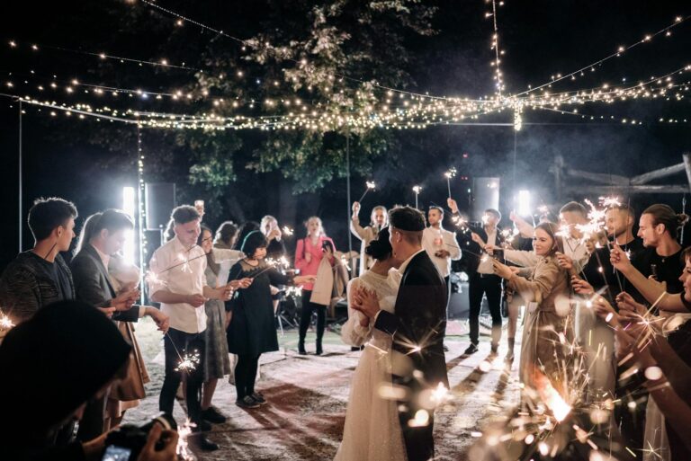 A joyful wedding reception with guests holding sparklers under string lights at night.