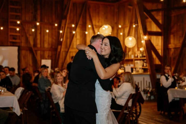 A couple sharing a dance at a wedding reception with guests seated at tables in a cozy, wooden barn.