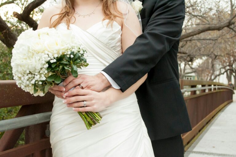 Bride holding a bouquet of white flowers, groom in a black suit embracing her.