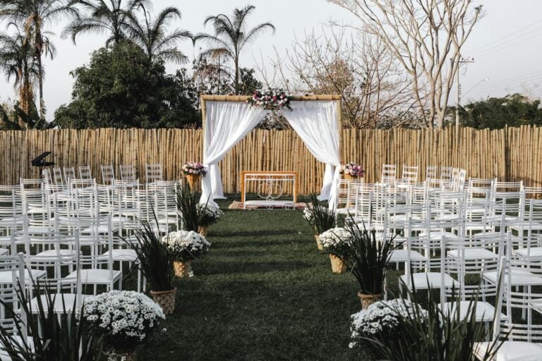 Outdoor wedding ceremony setup with white chairs, floral arch, and lush greenery.