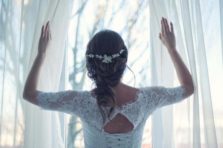 Bride gazing through sheer curtains in a wedding dress, enjoying the morning light.