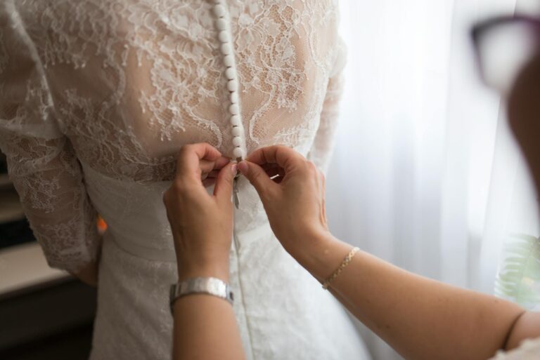 Elegant bride's dress being buttoned up, close-up shot of lace details and hands fastening the butto.