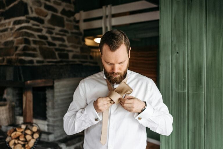 Groom preparing for his wedding, adjusting his beige tie in a cozy, rustic setting with stone and wo.